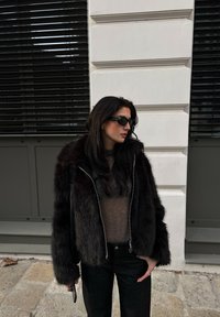 Faux fur-jacka i mörkbrunt med dragkedja fram och vida ärmar, i kombination med en ljusbrun topp och svarta jeans. Svarta solglasögon på.
