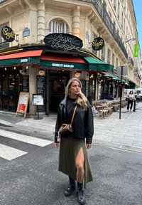 Musta nahkatakki, musta toppi, vihreä hame, jossa on reiden korkuinen halkio, maiharit ja pieni ruskea käsilaukku kahvilan edessä.