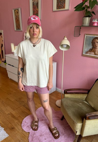 Camiseta oversized color beige claro, pantalones cortos a cuadros rojos, sandalias marrones con hebillas doradas, collares de plata y una gorra de béisbol rosa. Fondo rosa.