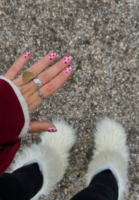 Main avec des ongles peints en rose, ornés de motifs de cœurs rouges. Anneaux en argent et bottes doublées de fourrure blanche sur une surface de gravier. Veste bordeaux visible.