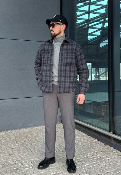 Chaqueta de camisa de cuadros gris oscuro sobre un cuello de tortuga gris claro, pantalones grises a medida y zapatos negros, combinados con una gorra negra y gafas de sol.
