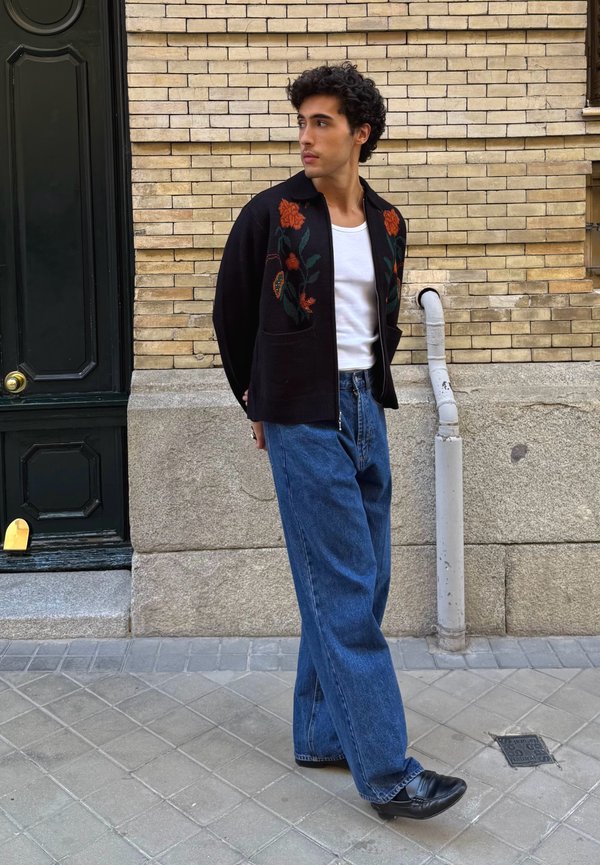 Black knit cardigan featuring floral embroidery, a white T-shirt, and blue wide-leg jeans. Black shoes complete the outfit. Set against an urban street backdrop.