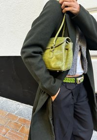 Small green croc-embossed leather handbag with a top handle and silver hardware, worn with a dark long coat and braided green belt.