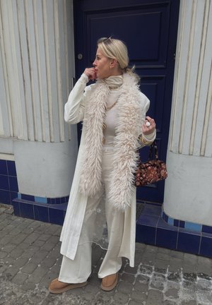 Mujer con conjunto en tonos crema de varias capas y bufanda larga de piel sintética, sosteniendo un bolso con estampado, de pie en una calle empedrada junto a una puerta azul.