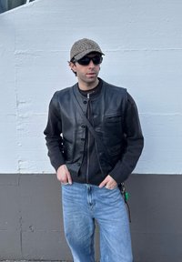 Μαύρη δερμάτινη γιλέκα πάνω από ένα σκούρο πουλόβερ, σε συνδυασμό με ανοιχτόχρωμα τζην. Τα αξεσουάρ περιλαμβάνουν καπέλο με λεοπάρ μοτίβο και γυαλιά ηλίου.