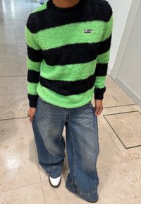 Henkilö, joka pukeutunut pörröiseen vihreän ja mustan raidalliseen neulepaitaan, väljät siniset farkut ja valkoiset kengät, seisoo sisätiloissa laattalattialla.