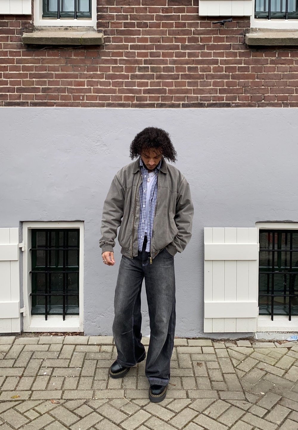 Szürke bomber dzseki egy kockás ing felett, laza sötét farmernadrággal és fekete cipővel párosítva. A háttérben téglafal és ablakok láthatók.