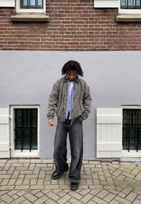 Szürke bomber dzseki egy kockás ing felett, laza sötét farmernadrággal és fekete cipővel párosítva. A háttérben téglafal és ablakok láthatók.