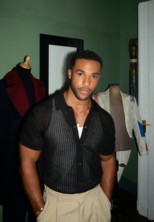 Camisa de manga corta de punto negro con un patrón de perlas, combinada con pantalones beiges; el fondo incluye un maniquí oscuro y una chaqueta clara.