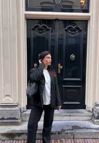 Blazer nero su una maglietta bianca, abbinato a pantaloni neri e una borsa a spalla, in piedi accanto a una porta nera ornata con hardware dorato.