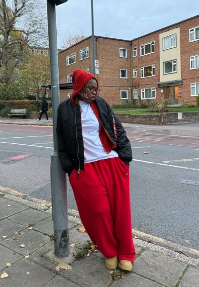 Chaqueta bomber negra sobre una camiseta blanca, pantalones deportivos rojos y zuecos amarillos. Capucha de punto roja cubre el cabello trenzado. De pie cerca de un poste en la calle.