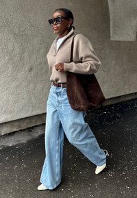 Γυναίκα που περπατά φορώντας μπεζ ζακέτα με φερμουάρ, ανοιχτό μπλε τζιν με φαρδιά πατζάκια, λευκά μυτερά παπούτσια, μεγάλα σκουρόχρωμα γυαλιά ηλίου και κρατώντας μία σκούρα καφέ σουέντ τσάντα.