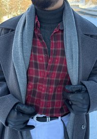 Manteau gris avec un col texturé, superposé à une chemise rouge à carreaux boutonnée. Des gants en cuir noir et un pantalon blanc complètent la tenue.