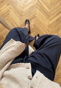 Cardigan bej tricotat cu nasturi din lemn, asortat cu pantaloni largi bleumarin și pantofi fara toc negri, pe un podea din lemn cu model în fir de pește.