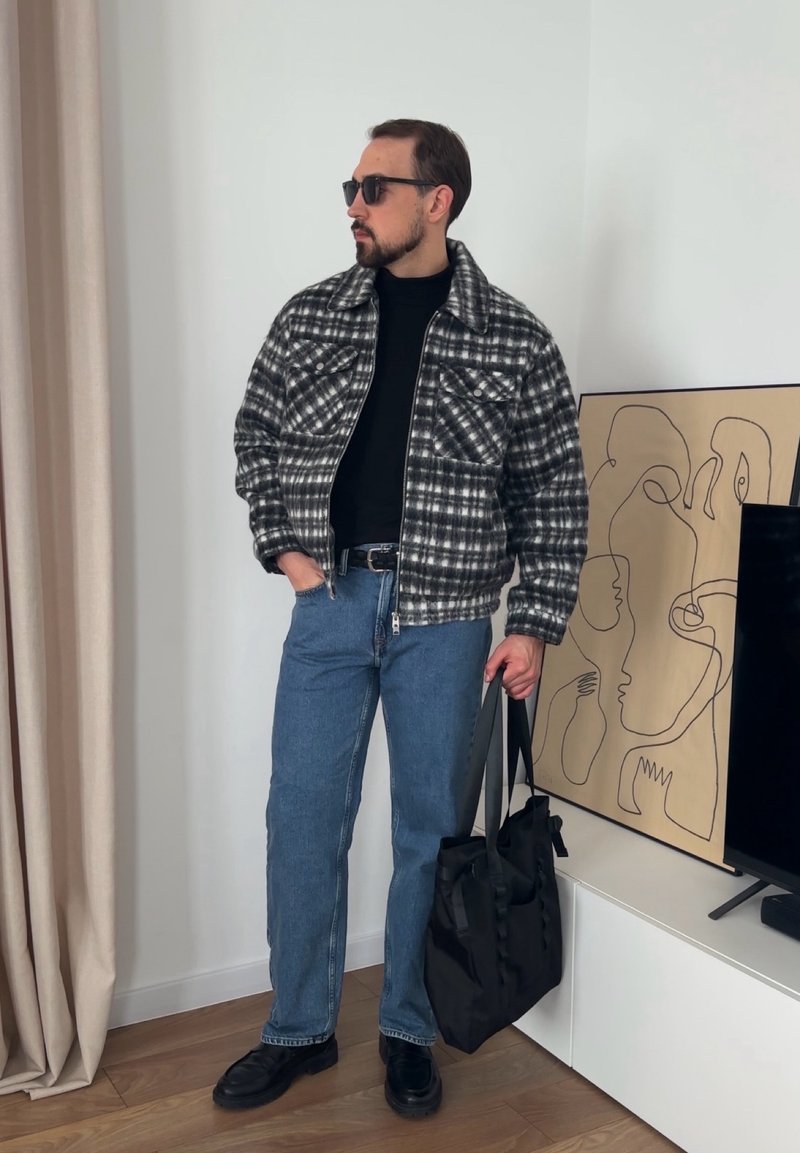 Veste à carreaux noir et blanc, col roulé noir, jean denim bleu clair, chaussures noires et sac fourre-tout noir. Fond uni et lisse.