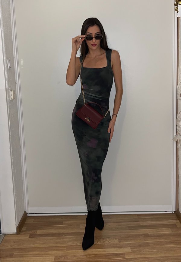 Vrouw in een mouwloze camouflagejurk, zwarte laarzen met hak, tilt met één hand haar zonnebril op en houdt een kleine bordeauxrode tas met een gouden kettingriem vast.