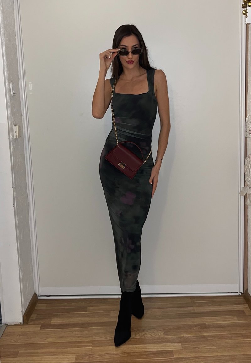 Femme en robe sans manches à motif camouflage, bottes noires à talons, levant des lunettes de soleil d'une main, tenant un petit sac bordeaux avec une bandoulière chaîne dorée.