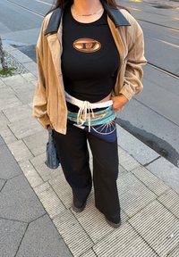 Haut noir à manches courtes avec logo orange, veste beige en velours côtelé, pantalon noir à jambes larges, et écharpe bleue à motif portée autour de la taille.