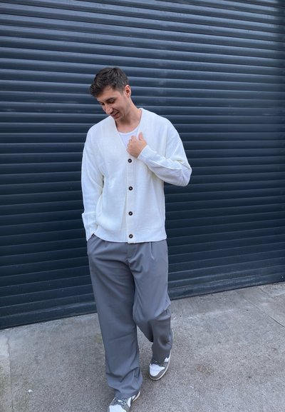 Cárdigan blanco con canalé y botones marrones, llevado sobre una camiseta ligera, combinado con pantalones grises holgados y zapatillas blancas. Fondo urbano.