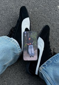 Oseba z belimi in črnimi športnimi copati ter svetlo modrimi kavbojkami drži telefon, na katerem je prikazan moški v črni jakni, kapi in širokih hlačah, ki stoji na prostem.
