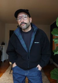 Mann med bart og briller iført svart caps, svart Carhartt-jakke, svart skjorte og blå jeans som står inne med hendene i lommene.