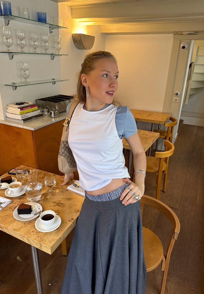 Giovane donna appoggiata a un tavolo del caffè con caffè, dessert, bicchieri e scaffali con libri dietro di lei.