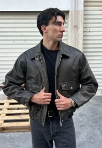 Veste en cuir vert foncé avec fermeture éclair à l'avant, poches à pression et taille réglable, portée par-dessus une chemise noire et un pantalon gris.