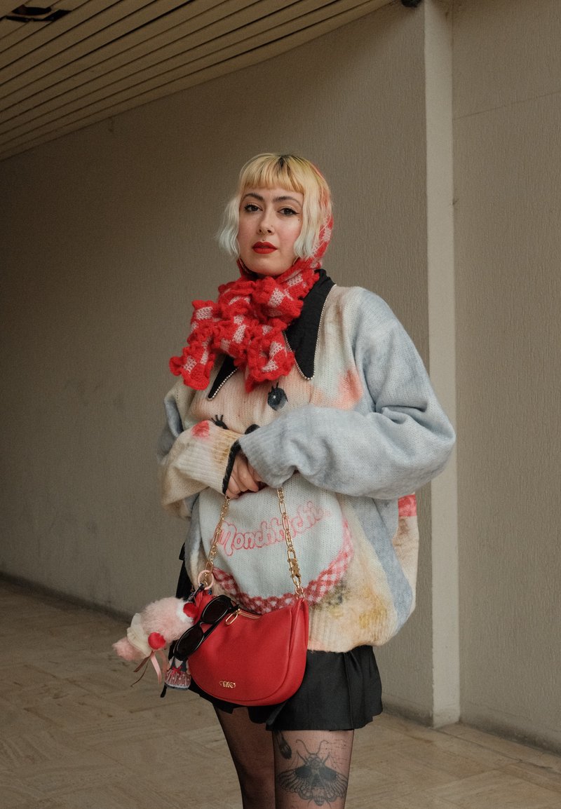 Oversized, gray sweater with colorful, soft textures and graphic design. Black skirt, red checkered scarf, and red handbag with gold chain.