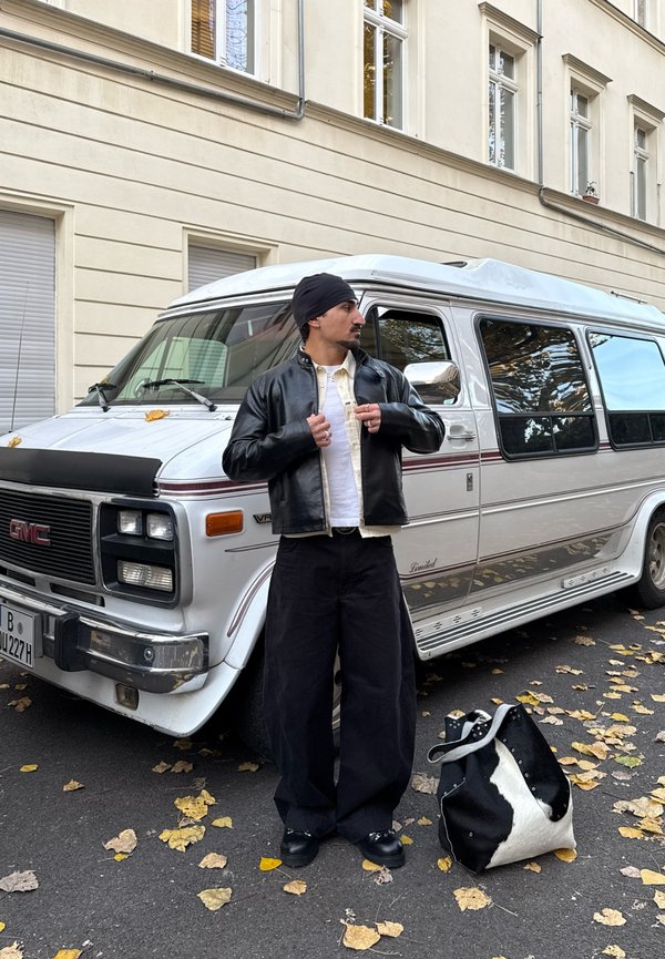 Black leather bomber jacket over a white shirt, loose black trousers, and a black beanie, next to a white GMC van and a patterned bag.