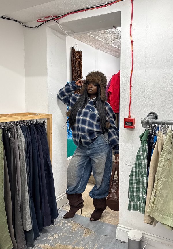 Femeie cu pălărie pufoasă, cămașă în carouri, blugi largi și cizme cu toc, pozând într-un magazin de haine între rafturi cu pantaloni și cămăși.