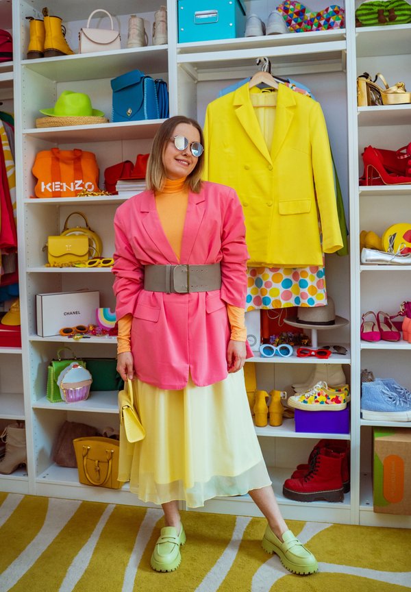 Kvinde iført pink blazer, orange rullekrave, gul nederdel og grønne sko står i et farverigt klædeskab med sko, tasker og farverige tøj.