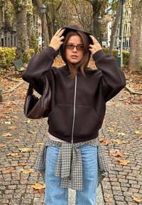 Juodas su užtrauktuku džemperis su dideliu gobtuvu, dėvimas ant šviesios palaidinės. Derinamas su laisvomis mėlynomis džinsomis ir languota marškinėlių, surištų aplink liemenį. Ruda rankinė.