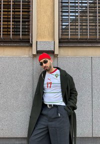 Άνδρας με γυαλιά ηλίου, κόκκινο σκουφί και φανέλα ποδοσφαίρου του Μαρόκου, στηρίζεται σε γκρι πέτρινο τοίχο με παράθυρα με κάγκελα, φορά σκούρο παλτό και ακουστικά.