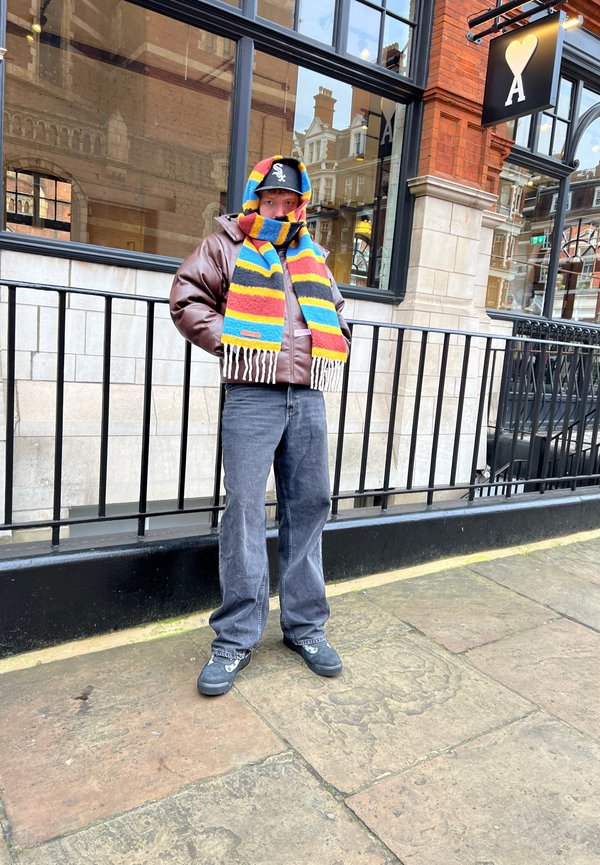 Chaqueta de plumas marrón, bufanda de rayas coloridas con flecos, jeans oscuros y zapatos negros; de pie frente a un escaparate de vidrio con ladrillo decorativo.