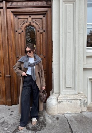 Abrigo largo marrón sobre un suéter gris, camisa blanca y pantalones a rayas. Bolso marrón y zapatillas grises. Fondo de puerta de madera.