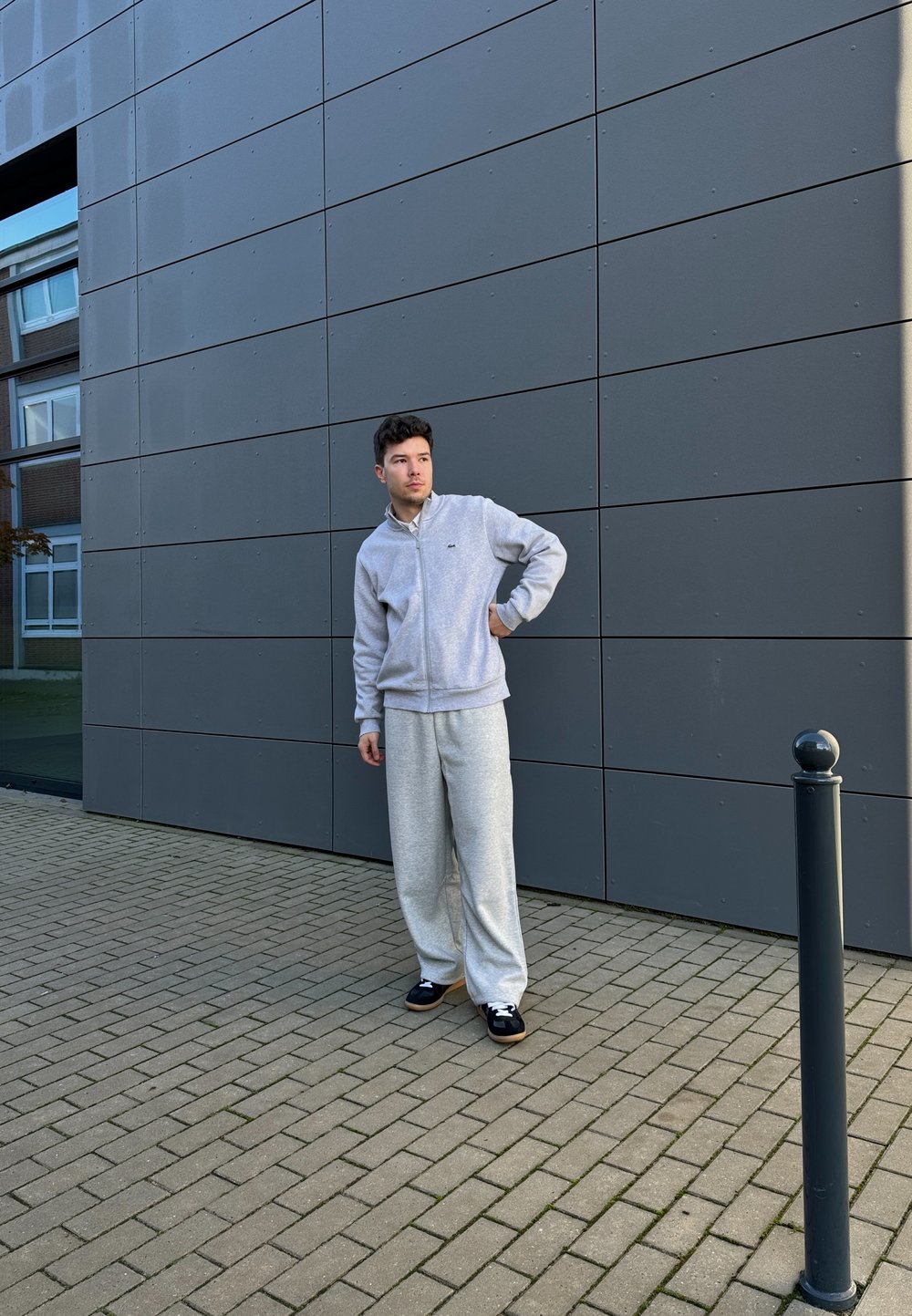 Ανοιχτό γκρι φούτερ με φερμουάρ και ταιριαστές φαρδιές φόρμες. Μαύρα παπούτσια με λευκές κορδόνες. Στηρίζεται σε μια σκοτεινή, μοντέρνα πρόσοψη κτηρίου.