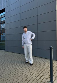 Ανοιχτό γκρι φούτερ με φερμουάρ και ταιριαστές φαρδιές φόρμες. Μαύρα παπούτσια με λευκές κορδόνες. Στηρίζεται σε μια σκοτεινή, μοντέρνα πρόσοψη κτηρίου.