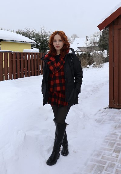 Mujer joven con cabello rojo que lleva una chaqueta negra, vestido de cuadros rojo y negro, y botas negras, de pie sobre un suelo cubierto de nieve cerca de una cerca de madera y edificios.