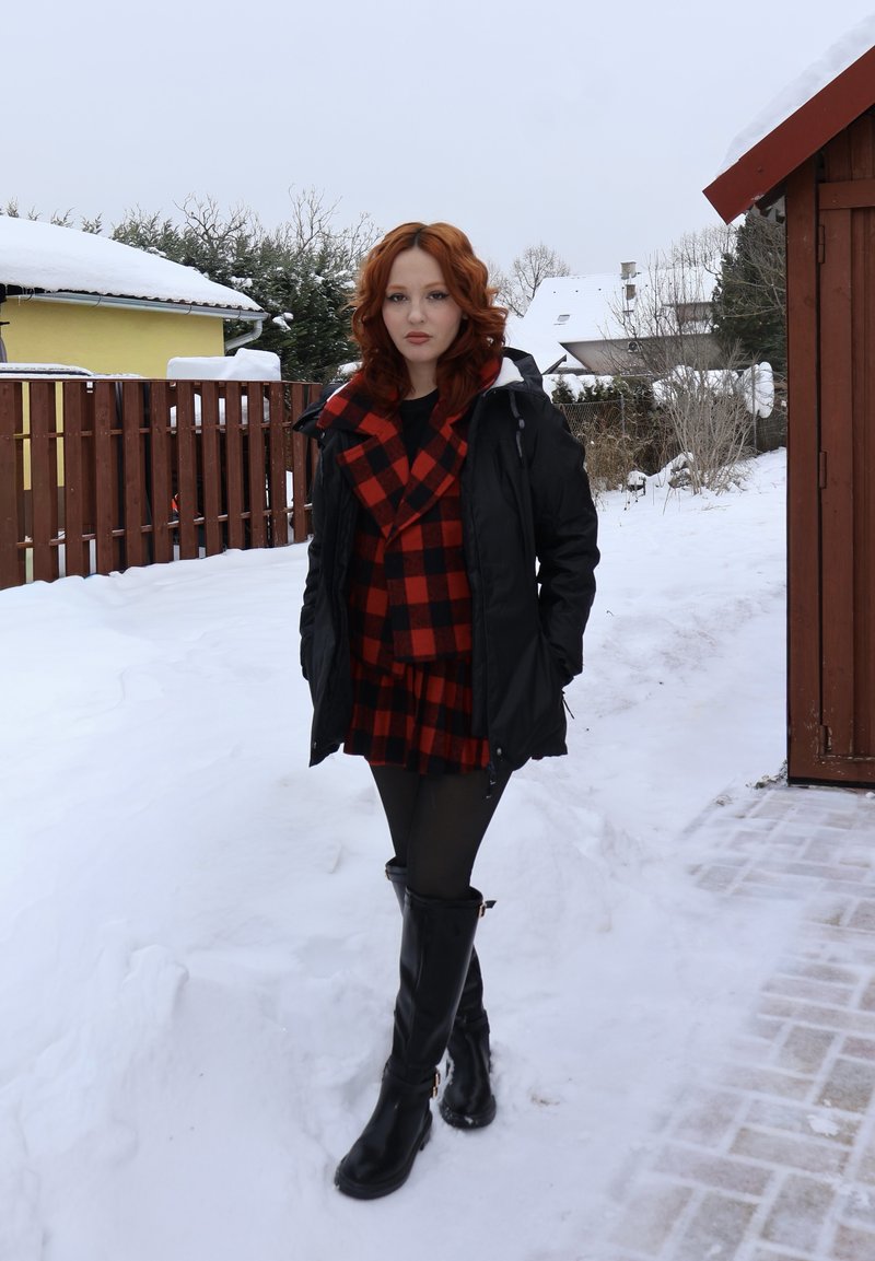 Jeune femme aux cheveux roux portant une veste noire, une robe à carreaux rouge et noir, et des bottes noires, debout sur un sol enneigé près d'une clôture en bois et de bâtiments.