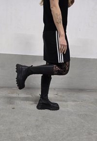 Sorte chunky støvler med en tyk sål, parret med en sort nederdel med hvide striber. Snørebåndsmønstrede leggings kan ses nedenunder.
