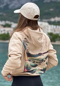 Béžová mikina s kapucí s potiskem malebné krajiny v modré, zelené a červené barvě na zadní straně. Obsahuje stahovací kapuci a žebrované manžety.