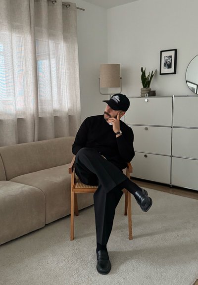 Una figura sentada con un suéter y pantalones negros, y zapatos negros, en una silla de madera tejida. De fondo hay cortinas beige, una lámpara y una cómoda.