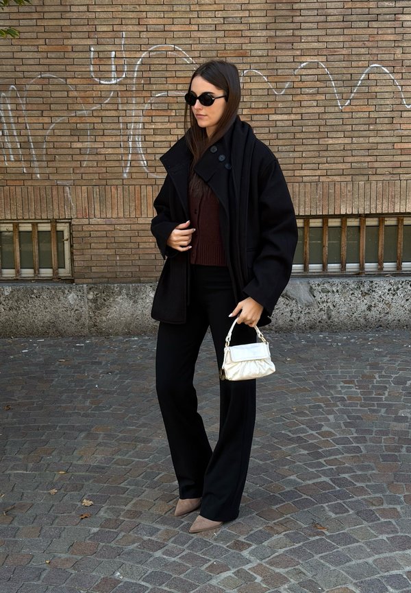 Femme portant un manteau noir, un pantalon noir, un pull marron, des lunettes de soleil, tenant un petit sac à main blanc, debout sur une rue pavée avec un mur en briques en arrière-plan.