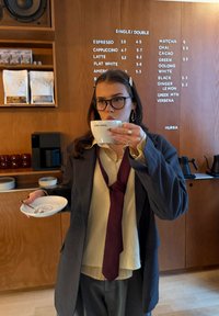 Femme portant des lunettes et une cravate lâche, qui boit un café dans une tasse blanche dans un café avec un tableau de menu en bois derrière elle.