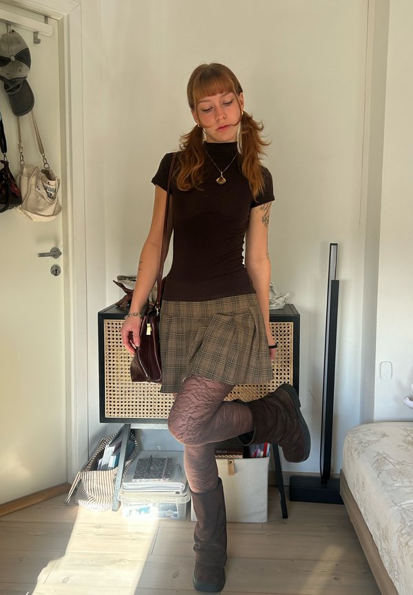 Una mujer posa en un dormitorio con un top negro, una falda de cuadros, medias estampadas, botas altas y un bolso al hombro.