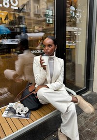 En sittende person i en hvit dress og lys grå turtleneck holder en kaffekopp. De sitter ved siden av en svart veske og et magasin på en trebenk.