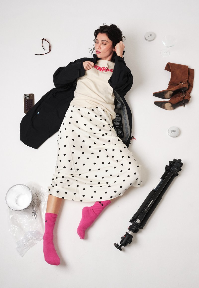 Femme allongée sur le sol portant une jupe à pois, une veste noire et des chaussettes roses, entourée de bottes marron, d'un trépied, d'un téléphone et de lunettes.