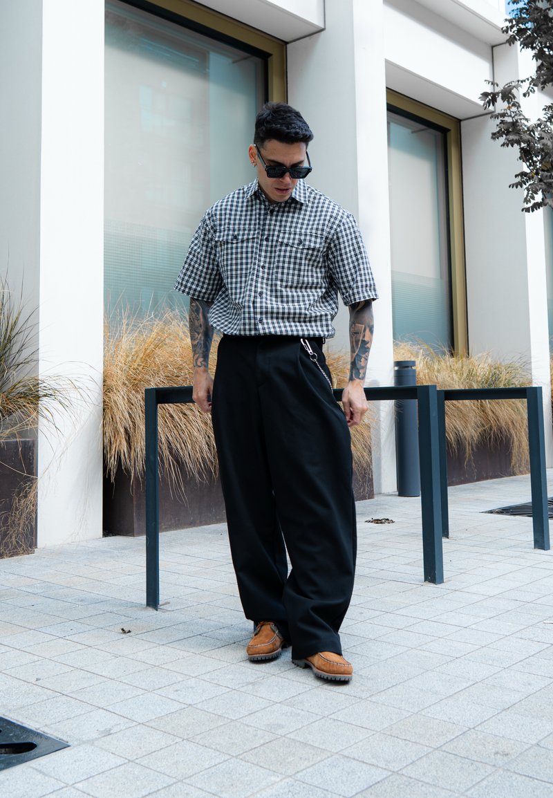 Camicia a maniche corte a scacchi bianco e nero, pantaloni larghi neri, mocassini marroni, occhiali da sole, in piedi davanti a un edificio moderno.