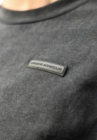 T-shirt de algodão preto com decote redondo, apresentando um logótipo "UNDER ARMOUR" em borracha preta elevado cosido na frente. Textura suave.
