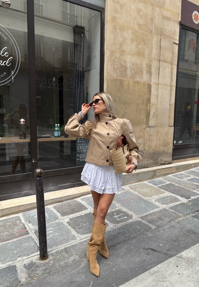Chaqueta de cuero marrón con mangas abullonadas, combinada con una minifalda blanca. Lleva botas altas de ante color beige y un bolso de mimbre. Gafas de sol oscuras.